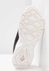 Semelle de sneaker Reebok noire et blanche montrant un motif de bande de roulement géométrique et le logo sur une surface blanche contre un fond blanc.