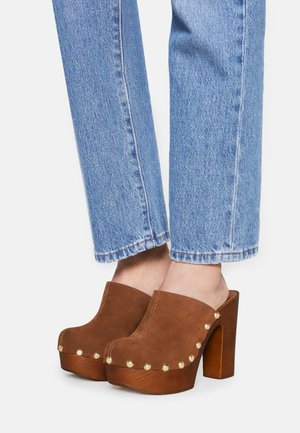 Sabots en suède brun à plateforme avec des clous dorés, portés avec un jean en denim bleu, montrant les pieds et les jambes inférieures sur fond blanc.