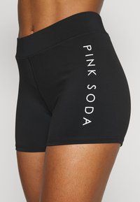 Černé sportovní šortky vyrobené z pružného materiálu, s širokým pasem a nápisem "PINK SODA" vytištěným bílou barvou podél strany.