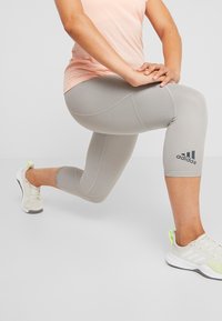 Harmaat capri-legginssit, joissa on teksturoidut paneelit ja Adidas-logo tummansinisellä. Yhdistettyinä valkoisiin ja neonvihreisiin urheilukenkiin sekä vaaleanpunaiseen puseroon.