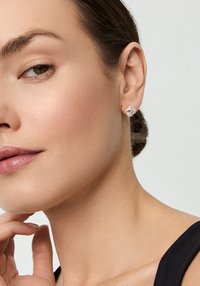 Różowe złote kolczyki-stud z kwadratowym wzorem, z centralnym przezroczystym kamieniem otoczonym mniejszymi kamieniami, osadzonymi na gładkiej tonacji skóry.