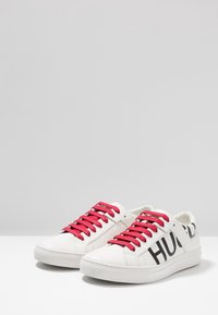 Baskets blanches avec des lacets rouges, marquage noir sur les côtés, en cuir lisse, bout rond et semelle plate en caoutchouc.