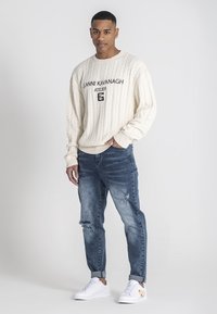 Weißer Kabelstrick-Pullover mit schwarzem Logo-Text, kombiniert mit ausgewaschenen blauen Jeans und weißen Sneakers mit sandfarbenen Akzenten. Lässiges Outfit.