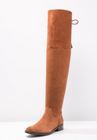 Bota alta de ante marrón, por encima de la rodilla, con suela plana y tirador trasero. Textura suave con detalles de costura mínimos a lo largo de las costuras.