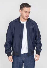 Marineblauer Bomberjacke mit einem Frontreißverschluss, gerippten Bündchen und Logostickerei. Aus glattem Stoff mit strukturierter Passform, kombiniert mit blauen Jeans.