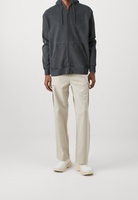 Felpa con zip grigia scura dotata di tasca anteriore, abbinata a pantaloni cargo beige chiaro con tasche laterali e sneakers color crema.