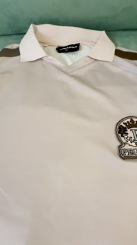 Šviesiai smėlinis polo marškinėliai su apykakle, su dekoratyviniu siuvinėtu ženklu kairėje pusėje ir tamsiomis juostomis ant pečių.