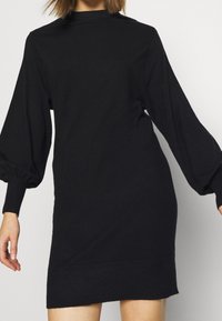 Robe noire à manches longues avec un col haut, un buste décontracté et des poignets côtelés, présentant un tissu texturé et une couleur unie.