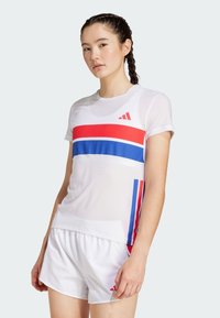 Camisa deportiva blanca con rayas horizontales rojas y azules, de manga corta, y logo de Adidas en rojo. Combinada con pantalones cortos blancos. Textura suave.