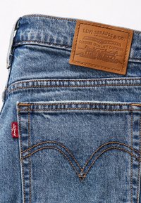 Poche arrière d’un jean en denim bleu avec des surpiqûres orange et un écusson en cuir marron Levi Strauss & Co. sur la ceinture.