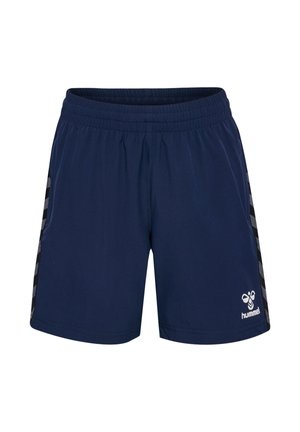 Jongen staat glimlachend in marineblauwe sportkleding met wit logo, zwarte sneakers en witte sokken tegen een effen lichte achtergrond.