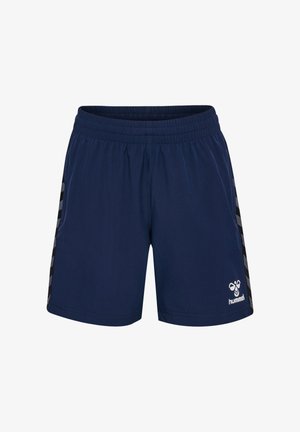 Niño sonriente de pie con ropa deportiva azul marino con logo blanco, zapatillas negras y calcetines blancos sobre un fondo claro y liso.