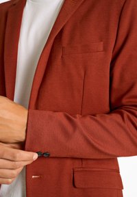 Blazer color ruggine con una vestibilità strutturata, tessuto testurizzato, taschino a petto singolo e chiusura con due bottoni sui polsini. Presenta cuciture a contrasto sottili.