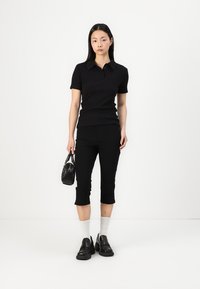Svart polo tröja i stickad tyg, tillsammans med svarta cropped byxor. Modellen håller en liten svart handväska och bär svarta skor med vita strumpor.