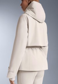 Svijetlo bež jakna s kapuljačom, labavog kroja, teksturirane površine i steznika u struku. Ima podesive manžete i moderan dizajn.