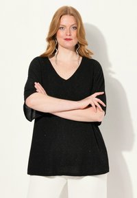 Černý top s krátkým rukávem a výstřihem do V, se třpytivou texturou a volným střihem. V kombinaci s bílými kalhotami pro kontrast.