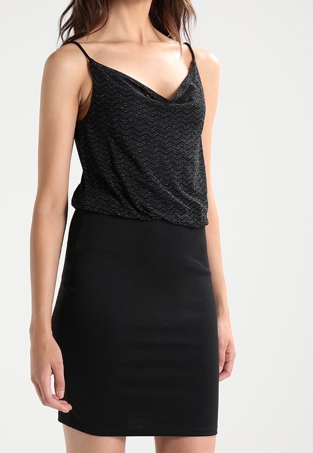Femme portant une robe noire sans manches avec un haut texturé scintillant et une jupe ajustée lisse sur un fond uni.