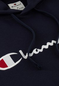 Marineblauwe hoodie met een witte geborduurde "Champion" logo met rode accenten. Zachte stof met een trekkoord op de capuchon en een zichtbaar label.