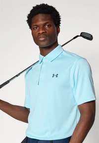 Lichtblaues poloshirt gemaakt van zachte, gestructureerde stof, met een drieknoopssluiting en een donker logo op de linkerborst.