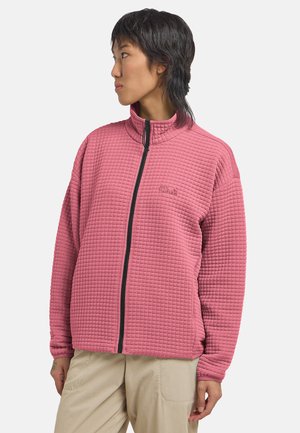 Personne tournée vers la gauche portant une veste matelassée rose zippée et un pantalon beige sur un fond uni.