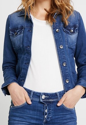 Une femme porte une veste en jean bleue et un jean par-dessus un haut blanc, les mains dans les poches.
