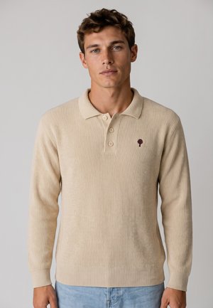 Jonge man met kort bruin haar, gekleed in een beige langärmelig gebreide polo met drie knopen en lichtblauwe spijkerbroek tegen een effen achtergrond.