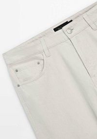 Section de la taille d’un jean denim écru avec poche avant, passants de ceinture et fermeture par bouton, montrant la texture du tissu et les détails de couture.