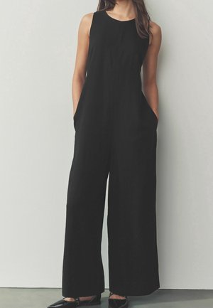 Femme portant une combinaison sans manches noire à jambes larges, les mains dans les poches, associée à des ballerines noires sur un sol gris.