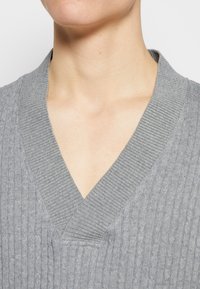 Suéter gris de punto con un escote en V acanalado y patrones texturizados en la tela, estrechamente ajustado en la zona del cuello.