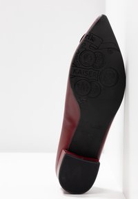 Semelle de chaussure en cuir rouge à bout pointu montrant des motifs de pièces embossés et le logo « KAISER » avec un embout de talon nervuré sur fond blanc.
