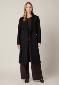 Czarny płaszcz dwurzędowy wykonany z teksturowanej tkaniny, z szerokimi klapami, dużymi guzikami oraz długą, swobodnie opadającą sylwetką. Połączony z ciemnobrązowym strojem.