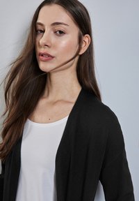Cardigan nero drappeggiato su una maglietta bianca. Texture liscia e design con apertura frontale. Lunghi capelli castani e mossi che incorniciano il viso. Sfondo neutro.