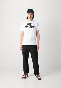 Hvid t-shirt med sort "JUST DO IT" logo, sorte cargo-bukser, mørk kasket og maroon- og hvide sneakers. Enkelt design, afslappet stil.