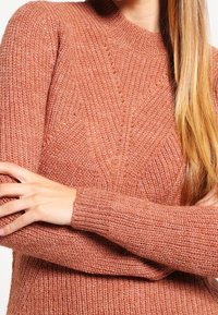 Pletený svetr v jemné barvě prašné růže, s vysokým výstřihem, strukturovanými vzory a žebrovanými manžetami. Materiál vypadá teplý a útulný.