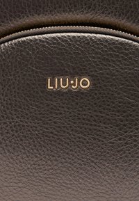 Nahaufnahme einer dunklen Ledertasche mit goldfarbenem "LIU·JO"-Logo, mit strukturiertem Oberflächen und einer Reißverschlusstasche oben.
