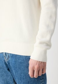 Main reposant sur le côté portant un pull blanc texturé et un jean bleu en denim sur un fond uni.