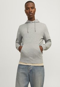 Gray hoodie with drawstring, front pocket, and ribbed hem. Worn over a light-colored layered shirt. Distressed blue jeans.