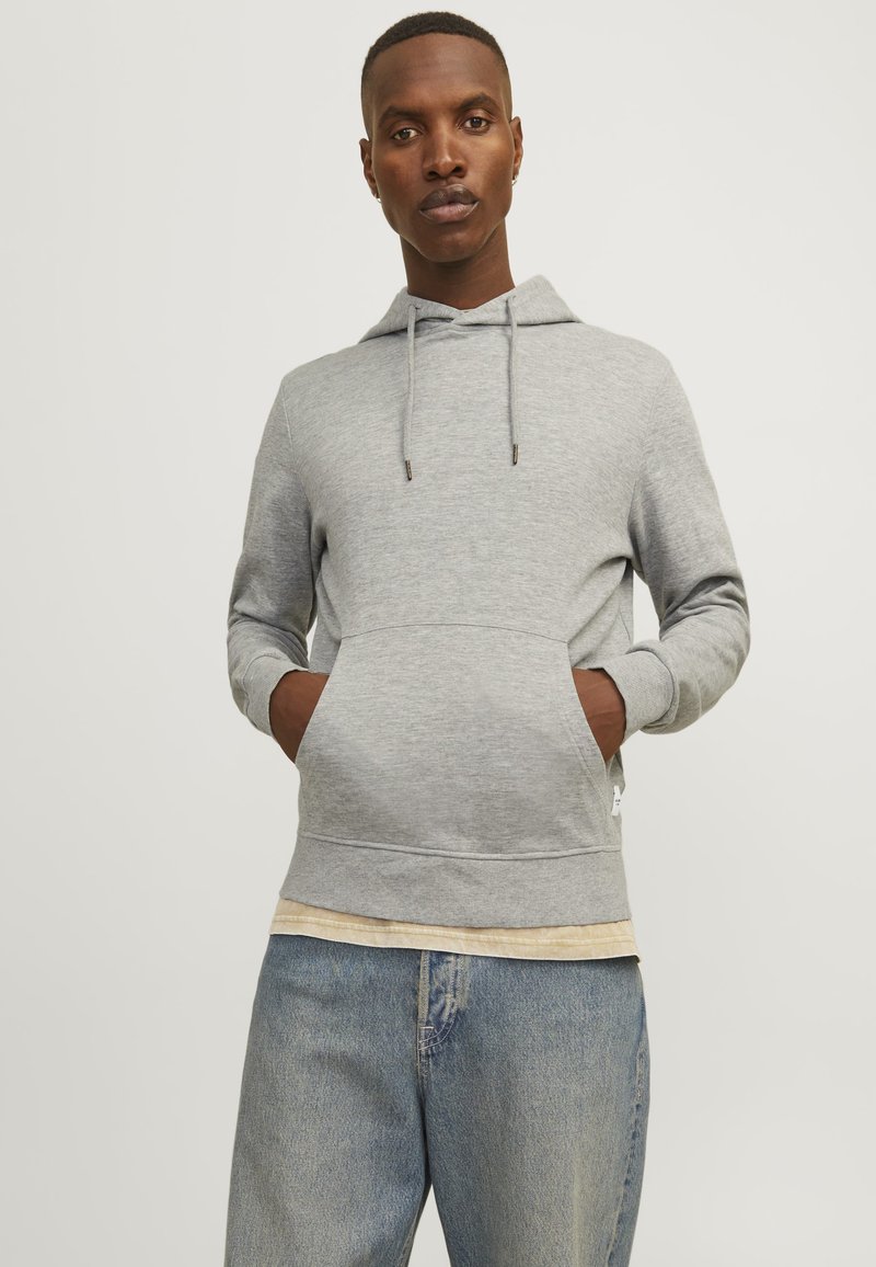 Gray hoodie with drawstring, front pocket, and ribbed hem. Worn over a light-colored layered shirt. Distressed blue jeans.
