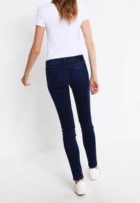 Vaqueros ajustados de color azul oscuro con cintura alta, que presentan una textura suave y costuras mínimas. Combinados con una camiseta blanca sencilla y zapatillas blancas.