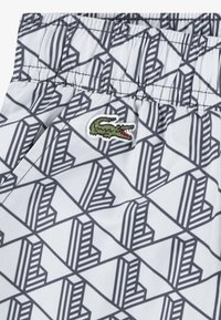 Σορτς με ελαστική μέση, κατασκευασμένα από λευκό ύφασμα με γεωμετρικό μαύρο σχέδιο. Περιλαμβάνουν πράσινο κεντητό λογότυπο κροκοδείλου.