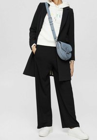 Person in schwarzem Mantel und Hose, weißem Hoodie mit buntem Text, weißen Sneakers und blauer Umhängetasche steht mit der Hand in der Tasche.