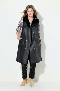 Gilet di pelle nera con colletto in pelliccia sintetica, dotato di bottoni dorati e due grandi tasche, abbinato a jeans scuri decorati e stivaletti nude alla caviglia.