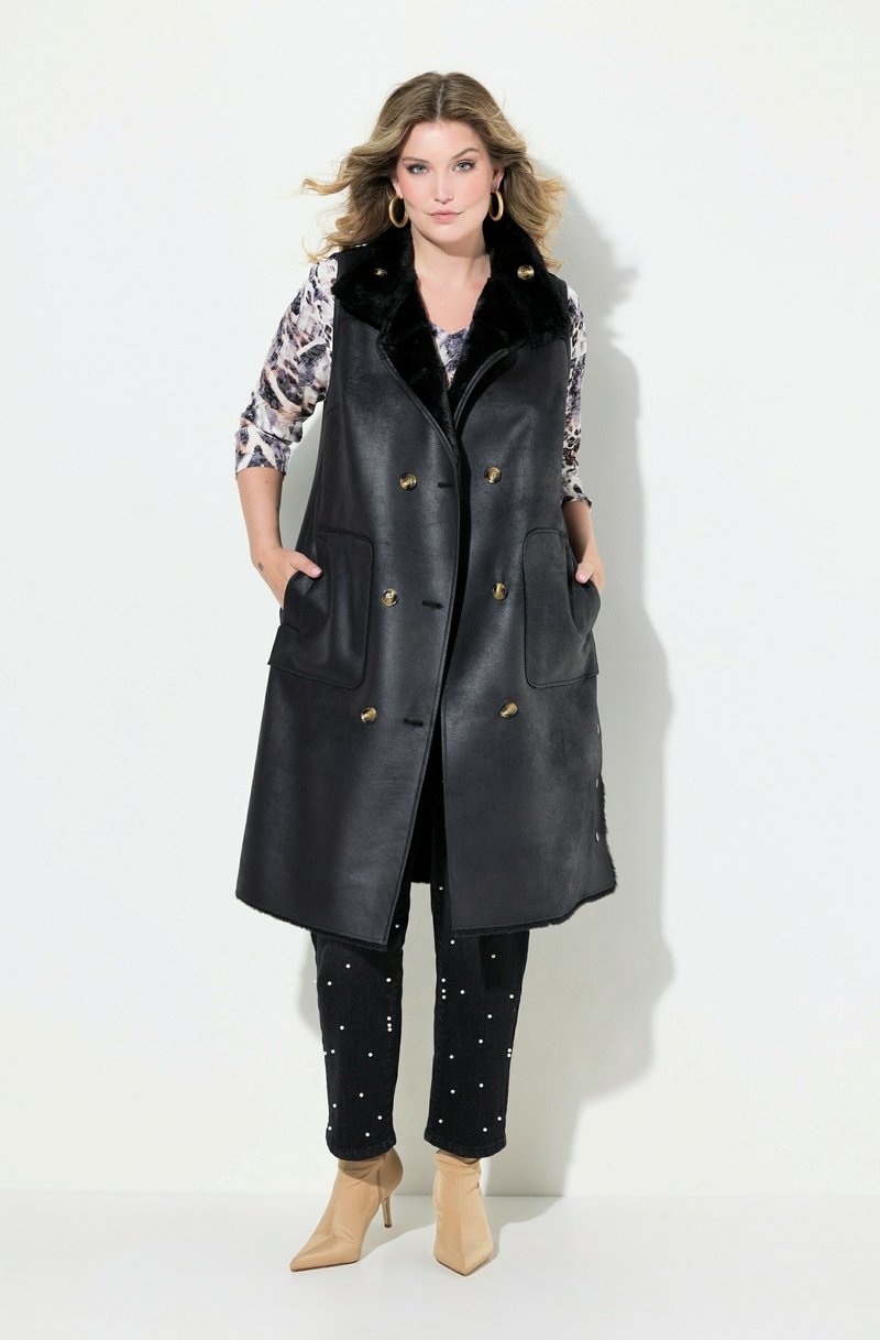 Gilet di pelle nera con colletto in pelliccia sintetica, dotato di bottoni dorati e due grandi tasche, abbinato a jeans scuri decorati e stivaletti nude alla caviglia.