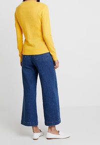 Gul tröja med långa ärmar, stickad textur, i kombination med vida mörkblå jeans med uppvikt nederkant, och vita slip-on skor.