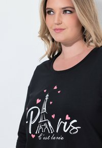 Schwarzes Langarmshirt mit einem weißen Eiffelturm-Grafik, pinken Herzen und dem Wort "Paris" in einer verspielten Schriftart. Weicher Stoff.