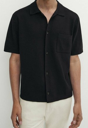 Homme portant une chemise noire à manches courtes avec poche poitrine, associée à un pantalon clair sur un fond uni.