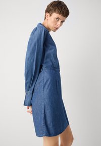 Denim obleka z dolgimi rokavi, oprijetim pasom in dolžino do kolen. Svetlo modra barva z gladko teksturo in diskretnimi šivi.