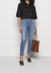 Camisa corta negra con cuello, jeans de tiro alto azul, sandalias planas negras con detalle anudado y un bolso tote marrón.