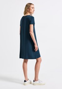 T-shirtklänning i mörk teal, knälång, kort ärm, avslappnad passform, slät textur. Matchad med vita sneakers med mörka accenter.