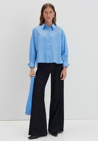 Camicia azzurro chiaro con bottoni e due tasche sul petto, abbinata a jeans a palazzo scuri e scarpe nere. Il modello tiene una borsa blu abbinata.